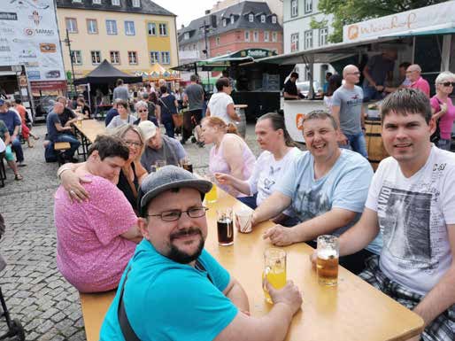 Bewohner und Mitarbeitende sitzen fröhlich an Biertischen auf einem Platz in der Saalfelder Altstadt, im Hintergrund Marktstände.