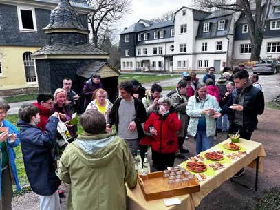 Gäste stehen um einen Tisch mit Gebäck und Getränken im Freien, dahinter das kleine Türmchen und die Häuser der Lebensgemeinschaft.
