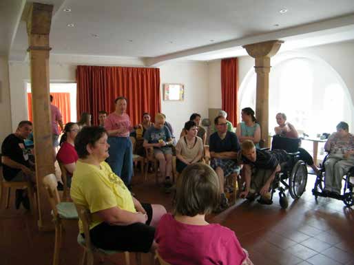 Viele Bewohnerinnen und Bewohner sitzen in der Cafeteria im Halbkreis auf Stühlen und in Rollstühlen, vorne eine Frau im gelben T-Shirt.