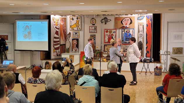 Preisverleihung in einem Saal voller bunter Kunstwerke: Auf der Bühne stehen mehrere Menschen, eine Frau erhält einen Preis, davor sitzt Publikum. Links eine Leinwand mit einer Präsentation.