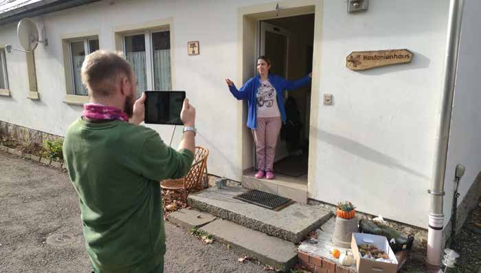 Ein Mann filmt mit einem iPad eine Bewohnerin, die mit ausgebreiteten Armen in der Tür des Kastanienhauses steht.