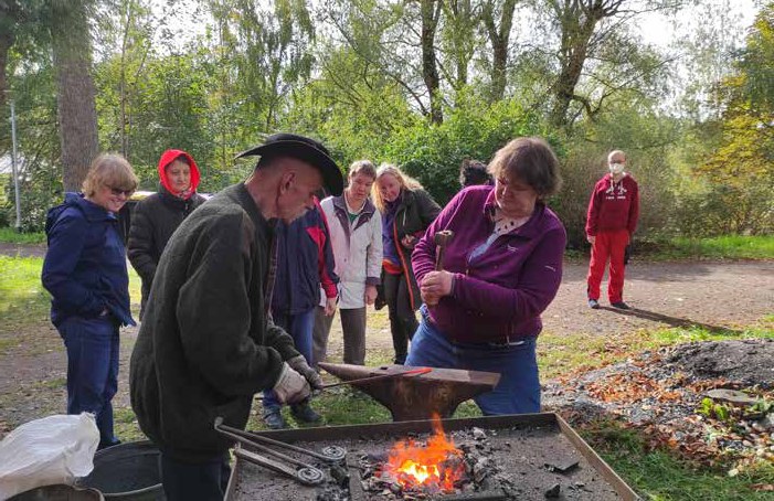 Im Freien glüht ein Schmiedefeuer neben einem Amboss, ein Schmied mit Hut hält ein Werkstück, eine Bewohnerin schlägt mit dem Hammer, weitere schauen zu.