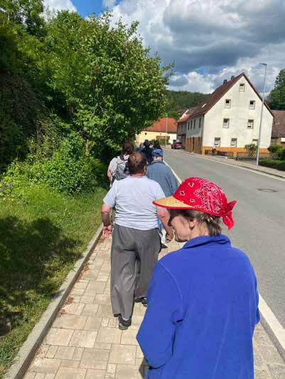 Die Gruppe spaziert auf einem Gehweg an einer Dorfstraße entlang, vorne eine Bewohnerin mit rotem Kopftuch und Hut.