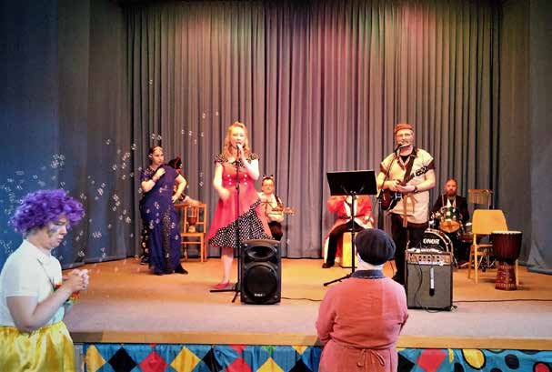 Auftritt auf der Bühne: eine Sängerin im roten Kleid, Musiker mit Gitarre und Trommeln, Seifenblasen in der Luft, vorne Zuschauer mit bunten Perücken.