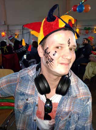Ein junger Mann mit Narrenkappe, aufgemalten Noten im Gesicht und Kopfhörern um den Hals lächelt in die Kamera.