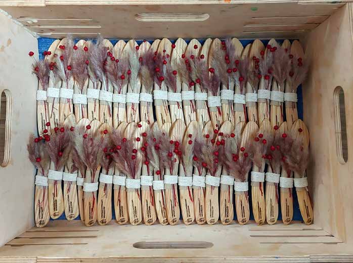Eine Holzkiste voller Geschenke: viele Buttermesser aus hellem Holz, je mit einem kleinen Strauß aus getrockneten Gräsern und roten Beeren, mit weißem Band gebündelt.
