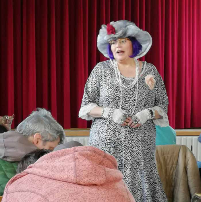 „Herta von der Bergbahn“ steht vor einem roten Bühnenvorhang: eine Frau mit grauem Hut mit roter Blume, lila Haaren, Perlenketten und leopardengemustertem Kleid. Vorne sitzen Zuschauer.