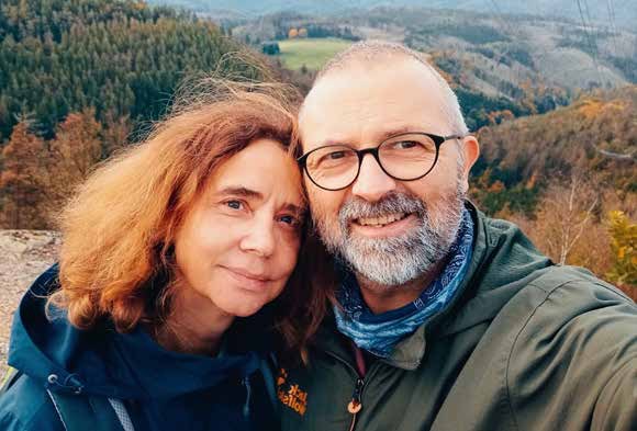 Selfie von Kathrin und Florian Naumann im Freien: eine Frau mit roten, lockigen Haaren und ein Mann mit grauem Bart und runder Brille lehnen die Köpfe aneinander, dahinter herbstliche, bewaldete Hügel.