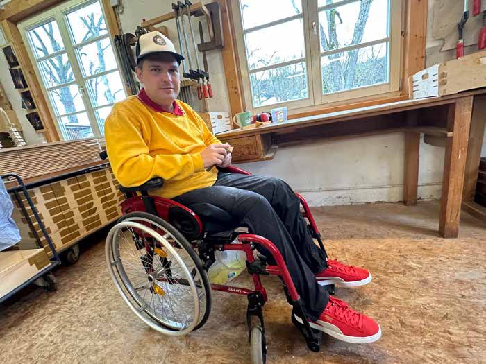 Paul sitzt in einem Rollstuhl in der hellen Schreinerei. Er trägt eine weiße Schirmmütze, einen gelben Pullover und rote Turnschuhe, hinter ihm Werkbänke und Fenster.