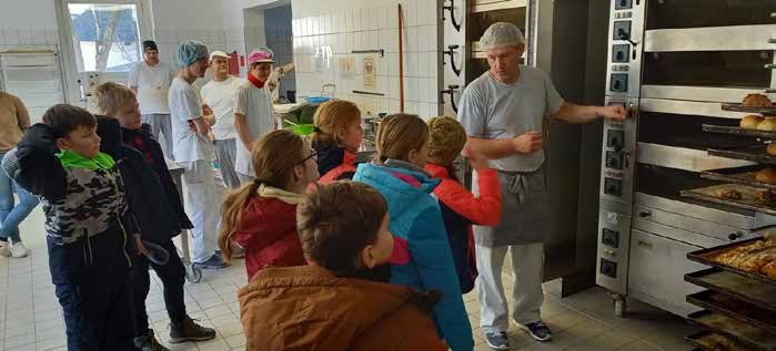 Eine Gruppe von Schulkindern in bunten Winterjacken steht in der Backstube. Ein Bäcker mit Haube zeigt ihnen den großen Backofen, dahinter weitere Mitarbeitende in weißer Kleidung.