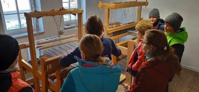 Schulkinder stehen in der Weberei um einen großen Webstuhl aus Holz.