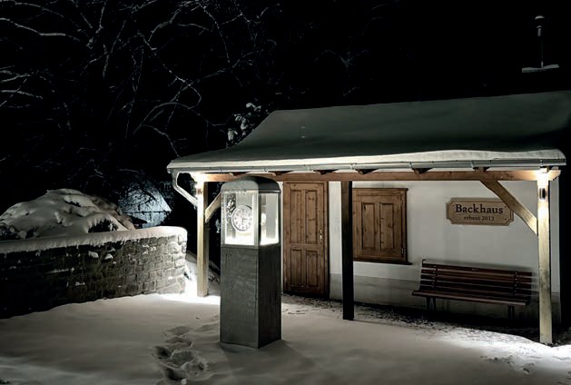 Das Backhaus nachts im Schnee: ein kleines Haus mit Holztür und Schild „Backhaus“ unter einem beleuchteten Vordach, davor eine Säule mit Uhr und Fußspuren im Schnee.