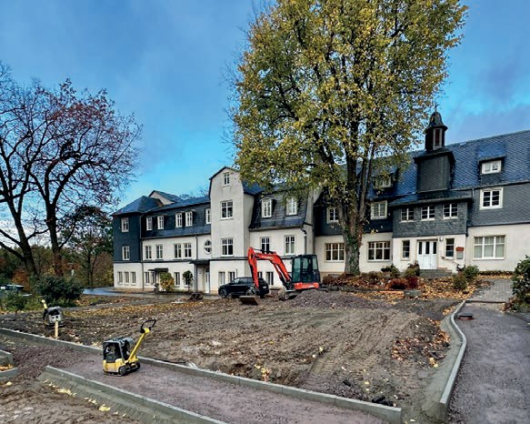 Der Platz während der Bauarbeiten: neue Wege, eine Rüttelplatte und ein roter Bagger vor den Häusern mit Schieferfassaden und einem großen Herbstbaum.