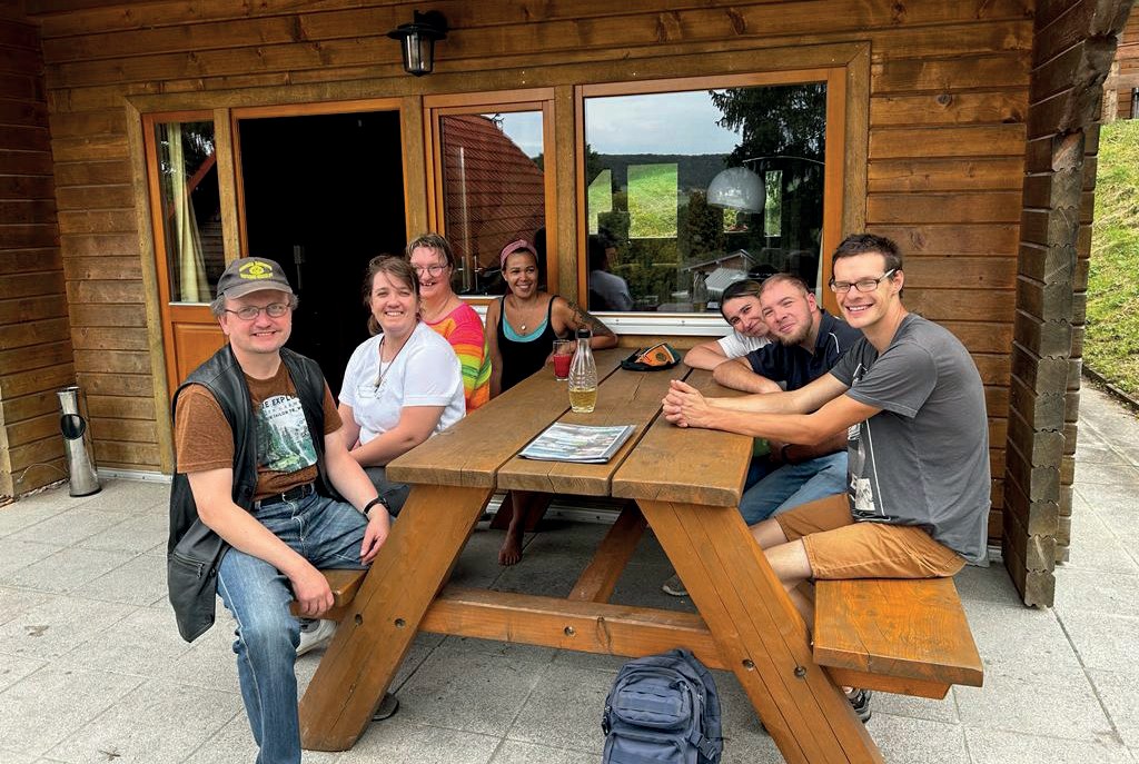 Die Gruppe sitzt fröhlich an einem Holztisch auf der Terrasse eines Holzbungalows.