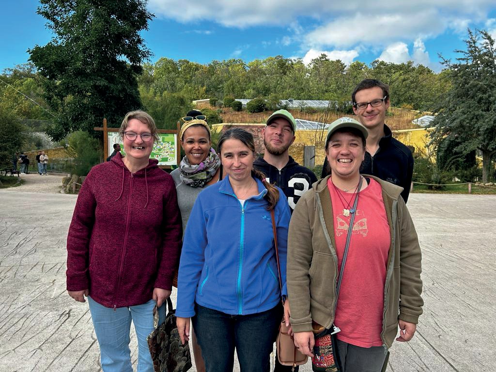 Sechs Urlauberinnen und Urlauber stehen lächelnd nebeneinander auf einem Platz im Zoopark, hinter ihnen Bäume und ein Gehege unter blauem Himmel.
