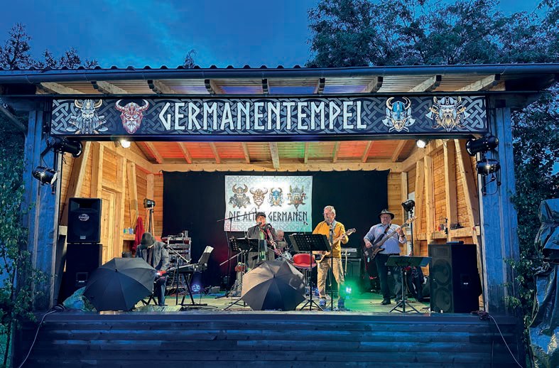 Die Freiluftbühne „Germanentempel“ am Abend: Die Band „Die Alten Germanen“ spielt im bunten Licht, vor der Bühne stehen aufgespannte Regenschirme.