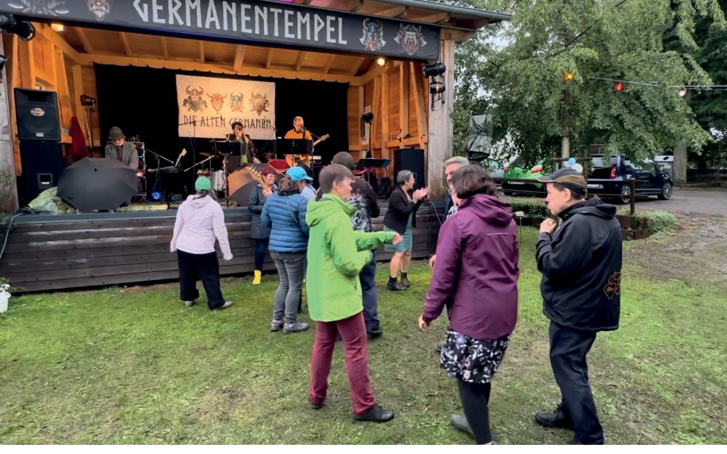 Gäste in Regenjacken tanzen auf der Wiese vor der Bühne „Germanentempel“, auf der die Band spielt.