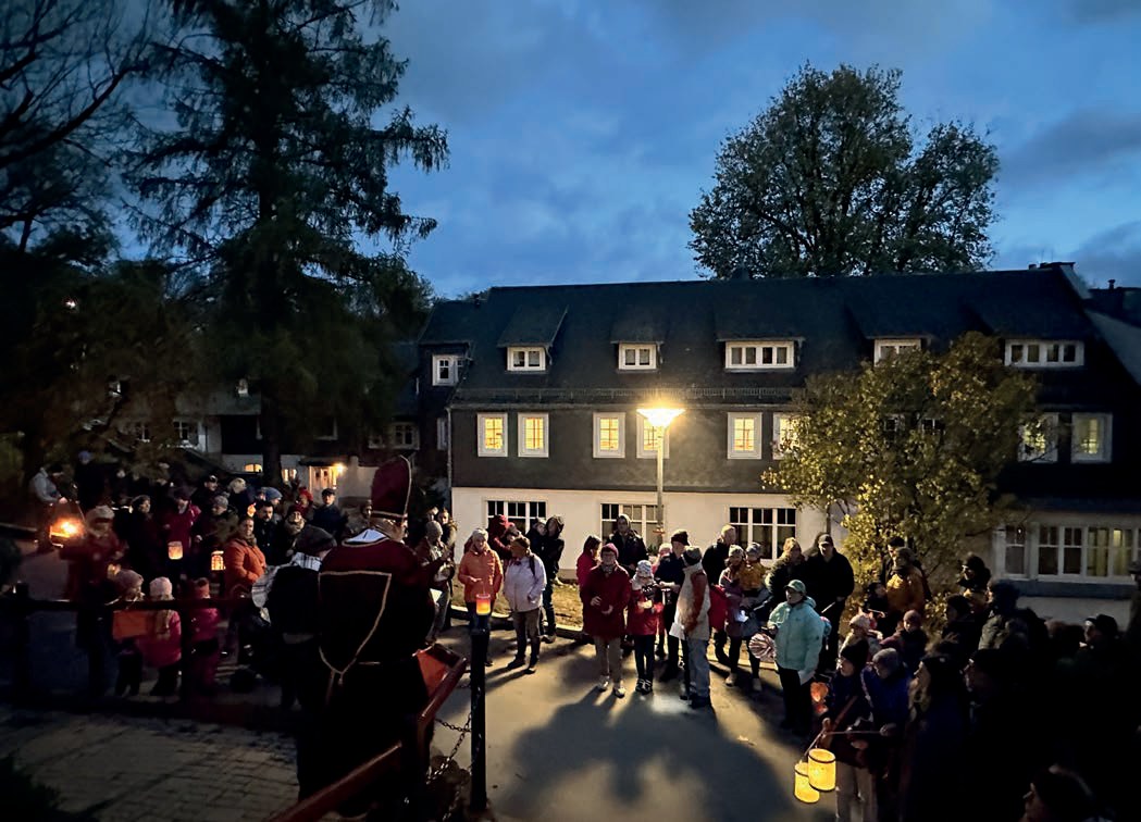 Laternenumzug in der Abenddämmerung: Viele Menschen mit leuchtenden Laternen stehen vor einem großen Haus mit erleuchteten Fenstern, vorne Sankt Martin in rotem Umhang.