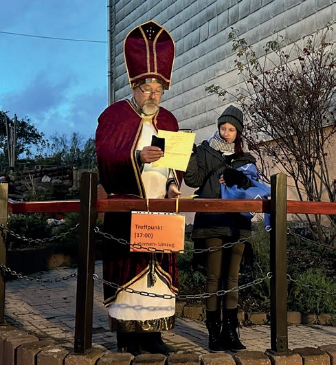 Sankt Martin mit Bischofsmütze und rotem Umhang liest am Abend von einem Zettel vor, an seinem Pult hängt ein Schild „Treffpunkt 17:00 Laternen-Umzug“.