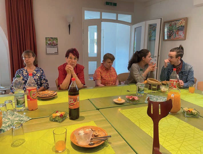 Weitere Gäste sitzen an der Tafel unter einem großen Fenster mit Rundbogen.