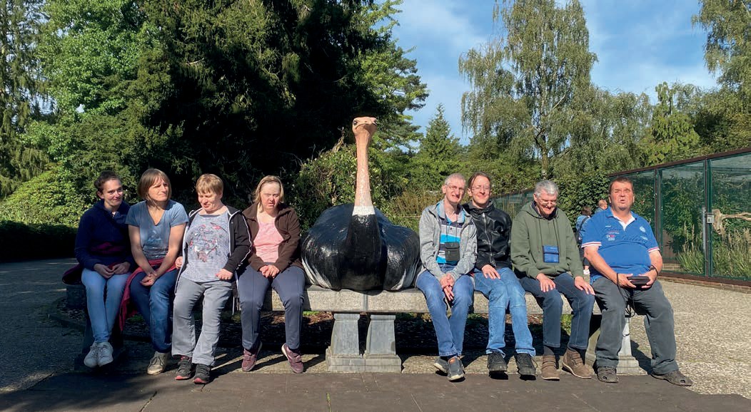 Die Urlaubsgruppe des Morgensternhauses sitzt in der Sonne nebeneinander auf einer langen Steinbank, in der Mitte steht die Figur eines großen Straußes.