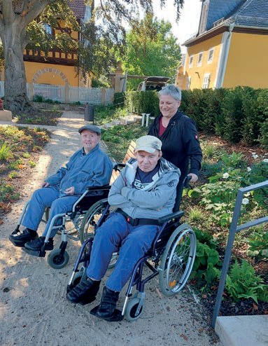 Zwei ältere Herren in Rollstühlen auf einem Gartenweg in der Sonne, hinter ihnen eine Mitarbeiterin.