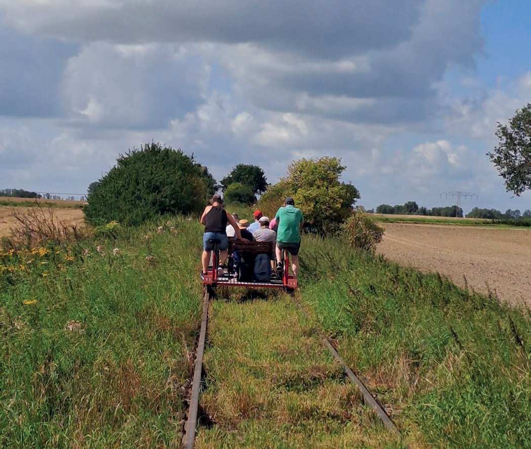 Eine Draisine mit mehreren Menschen fährt auf einem alten, mit Gras bewachsenen Gleis zwischen Feldern und Büschen in die Ferne, darüber ein weiter Himmel mit Wolken.