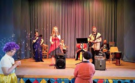 Auftritt auf der Bühne vor einem Vorhang: Eine Sängerin im roten Kleid singt, daneben Musiker mit Gitarre und Trommeln, vorne Zuschauer in Faschingskostümen.