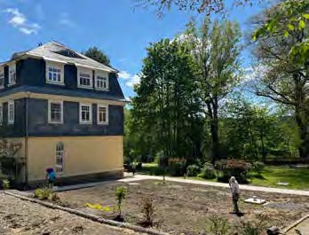 Ein Haus mit Schieferfassade und hellem Erdgeschoss, davor eine frisch bepflanzte Fläche, auf der jemand arbeitet.
