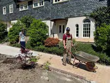 Zwei Gärtner bei der Arbeit vor einem Haus: einer mit einer Schubkarre, einer mit einer Harke an einem frisch bepflanzten Beet.