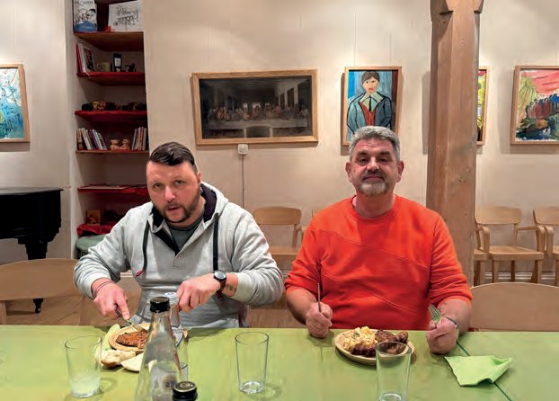 Zwei Männer sitzen beim Abendessen am Tisch, vor sich Teller mit Gegrilltem und Kartoffelsalat.