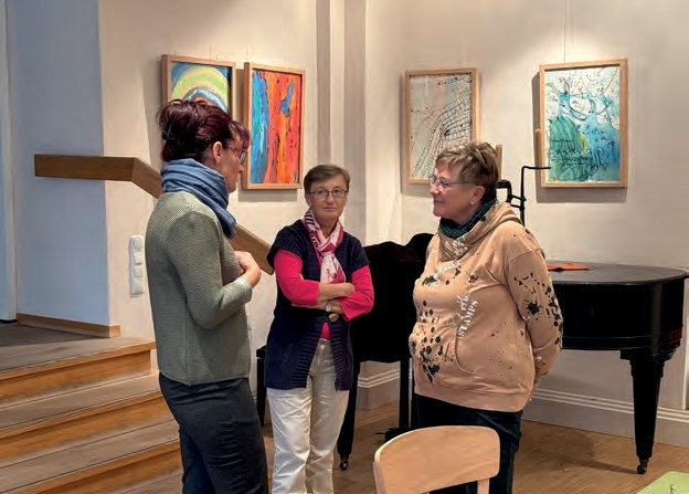 Drei Frauen unterhalten sich in der Cafeteria vor einem Flügel und bunten Bildern an der Wand.