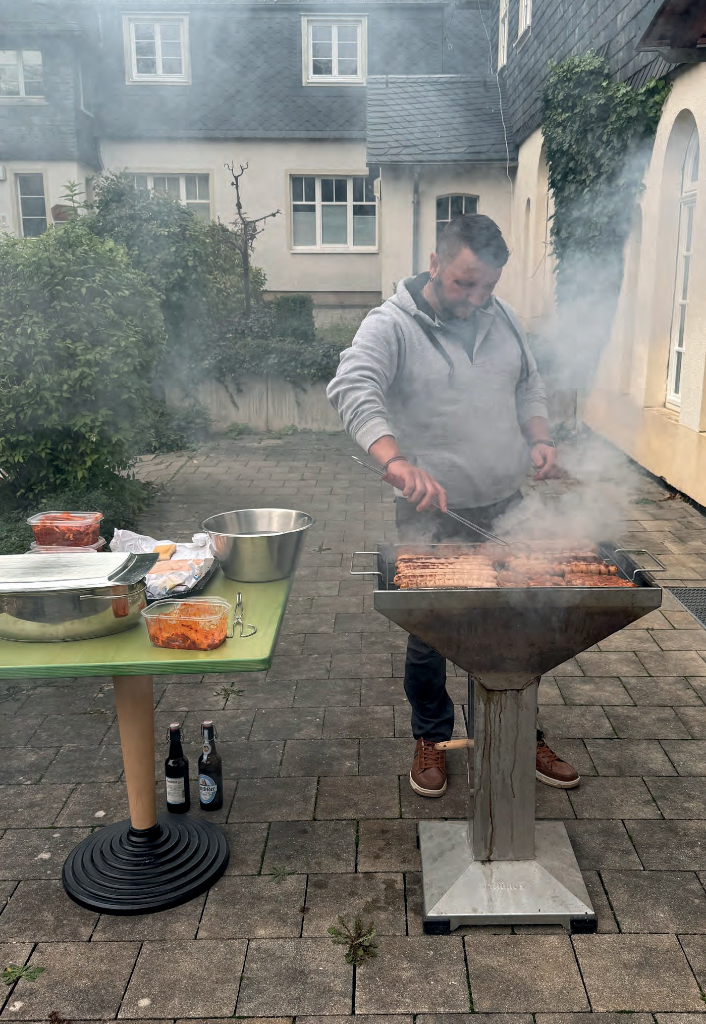 Ein Mann grillt im Hof Fleisch auf einem großen Feuerkorb-Grill, Rauch steigt auf. Daneben ein Tisch mit Schüsseln.