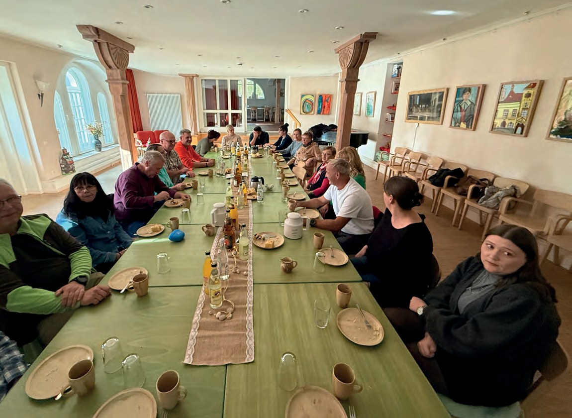Eine lange, festlich gedeckte Kaffeetafel in der Cafeteria mit grünen Tischen, Tellern und Tassen. An beiden Seiten sitzen Bewohner, Angehörige und Mitarbeitende, an den Wänden hängen bunte Bilder.