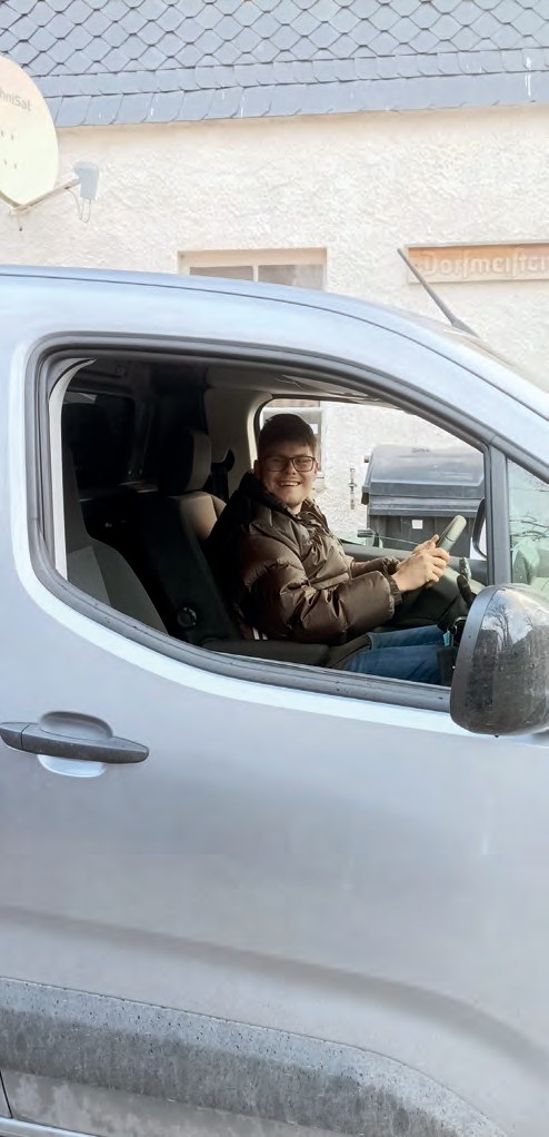 Dominic sitzt am Steuer eines silbernen Lieferwagens, die Fahrertür ist offen, er lächelt in die Kamera.
