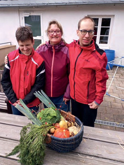 Drei Bewohner in roten Jacken stehen lächelnd hinter einem Holztisch, auf dem ein Korb voller Gemüse steht: Lauch, Kohl, Kürbis, Möhren und Kräuter.