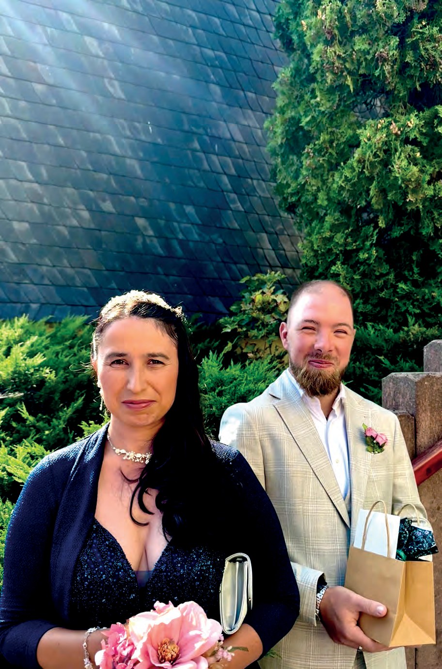 Das Brautpaar im Freien: Sandra in einem dunkelblauen Kleid mit einem rosa Blumenstrauß, Sven mit Bart im hellen karierten Anzug mit Ansteckblume, er hält eine Geschenktüte.