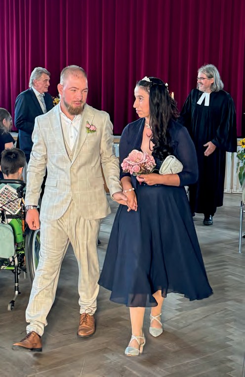 Das Brautpaar geht Hand in Hand durch die Festhalle, Sandra im dunkelblauen Kleid mit Blumenstrauß, Sven im hellen Anzug. Hinter ihnen ein Pfarrer im Talar und Gäste vor dem roten Bühnenvorhang.