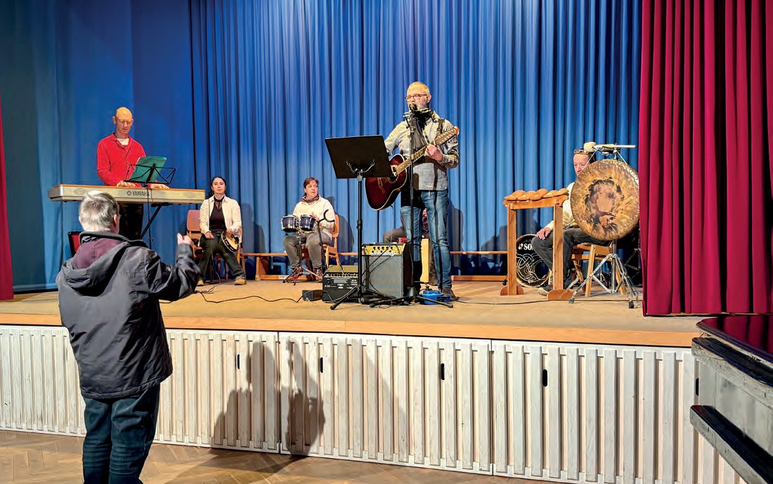 Musikerinnen und Musiker auf der Bühne der Festhalle vor einem blauen Vorhang, mit Keyboard, Gitarren und einer großen Trommel. Vorne steht ein Zuschauer und fotografiert.