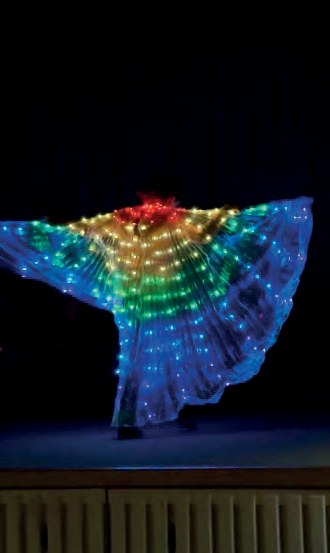 Auf einer dunklen Bühne breitet eine Tänzerin weite Flügel aus, die mit Lichtern in allen Regenbogenfarben leuchten.