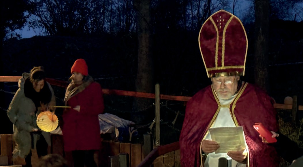 Abends im Dunkeln: Sankt Martin mit roter Bischofsmütze und rotem Umhang liest von einem Blatt vor. Neben ihm stehen Menschen mit leuchtenden Laternen.