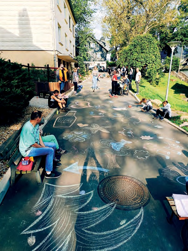 Die neue Straße zwischen den Häusern ist über die ganze Breite mit bunten Kreidebildern bemalt: Blumen, Kreise, Wellen und ein großer Baum. Ein Mann sitzt am Rand auf einer Bank, weiter hinten malen andere.