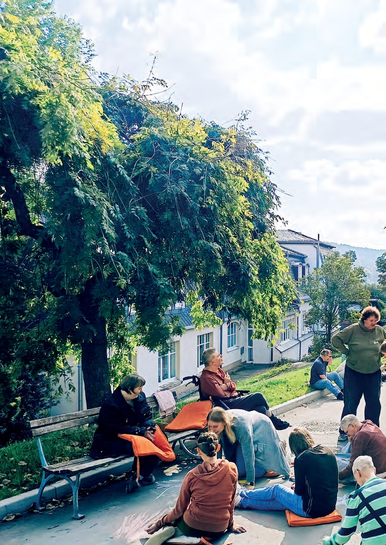 Bewohnerinnen und Bewohner sitzen auf Kissen auf der Straße und malen mit Kreide, über ihnen ein großer Baum, im Hintergrund Häuser.