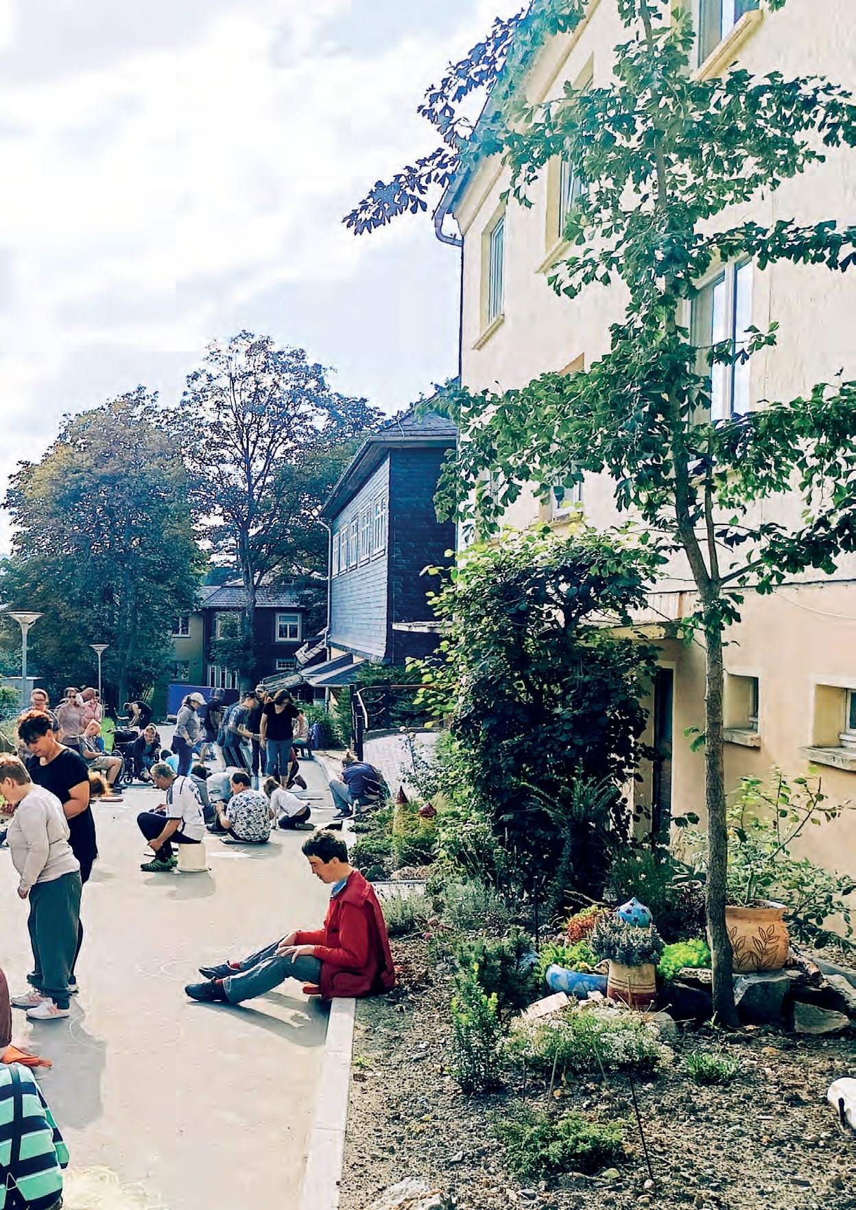 Blick die Straße entlang: Viele Menschen knien und sitzen auf dem Asphalt und malen, rechts ein frisch bepflanztes Beet vor einem Haus.