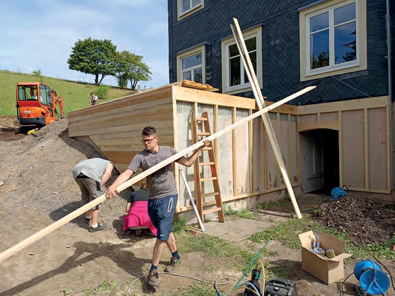 Bauarbeiten an der Terrasse: Zwei Männer tragen ein langes Kantholz, an der Hauswand steht schon die neue Holzverschalung, im Hintergrund ein kleiner Bagger am Hang.