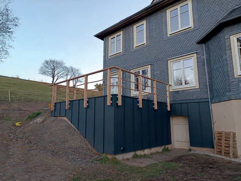 Die neue Terrasse am August-Halm-Haus: ein erhöhter Unterbau, verkleidet mit dunkelgrauem Blech, oben ein Geländer aus hellem Holz. Daneben das Haus mit Schieferfassade, davor frisch aufgeschüttete Erde.