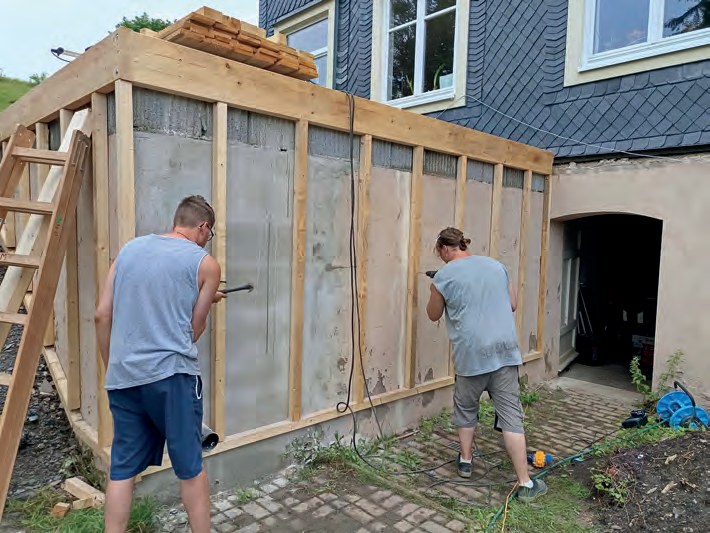 Zwei Männer arbeiten mit Werkzeug an der Holzverschalung des Terrassenunterbaus, links lehnt eine Leiter.