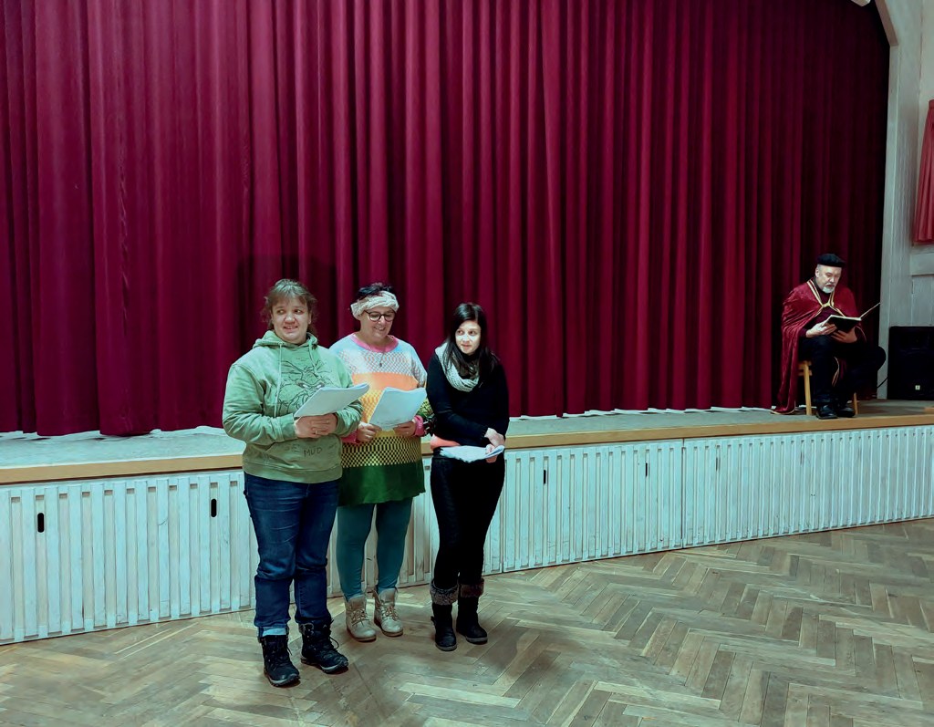 Drei Frauen stehen mit Textblättern vor dem roten Bühnenvorhang in der Festhalle, rechts am Bühnenrand sitzt ein Mann in rotem Umhang.