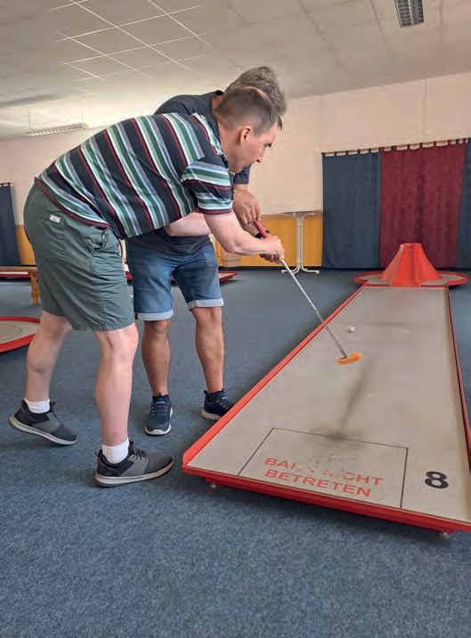 Ein Mann im gestreiften Poloshirt schlägt beim Minigolf in einer Halle den Ball, hinter ihm ein zweiter Spieler.