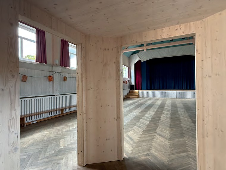 Blick durch einen neuen Eingang aus hellem Holz in die Festhalle mit Fischgrätparkett, Bänken an der Wand und der Bühne mit dunkelblauem Vorhang.